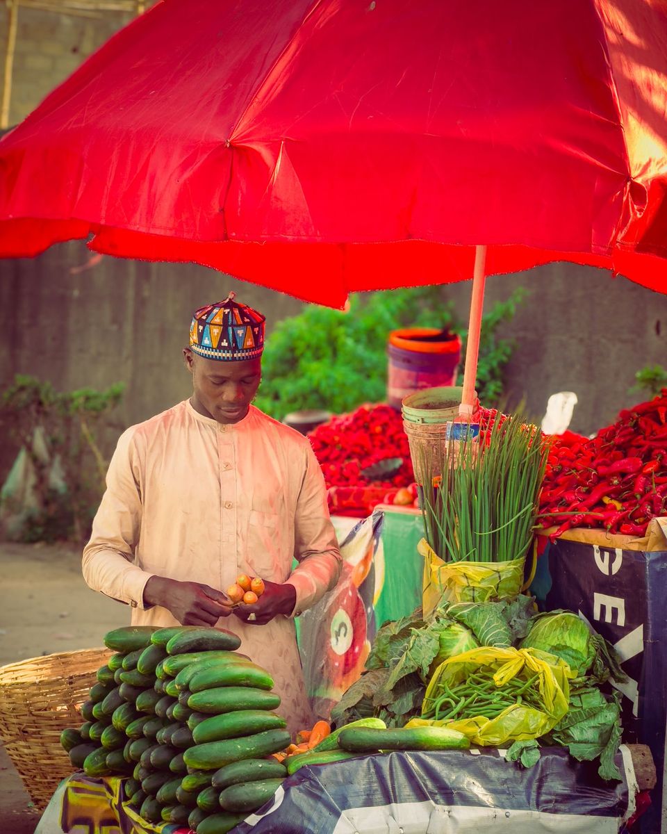 A vendor arranging cucumbers, peppers and greens at a market stall under a red umbrella in Lagos